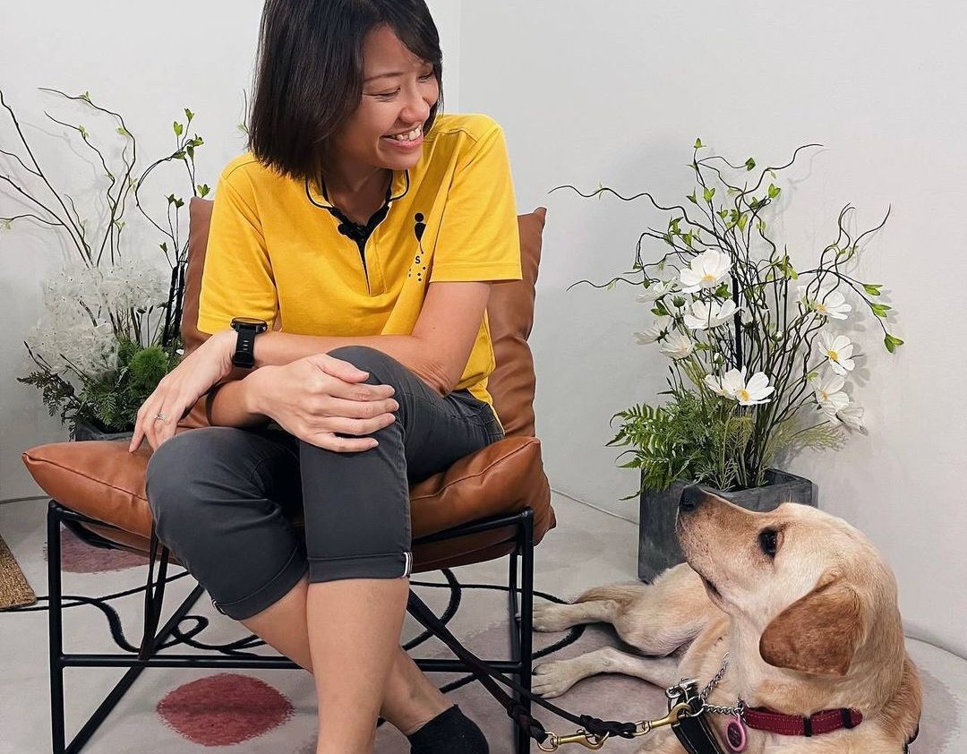 Guide Dog Mobility Instructor Christina is sitting on a brown chair and looking at guide dog Orinda, who is lying down beside the chair. Guide dog Orinda is looking back at Christina.