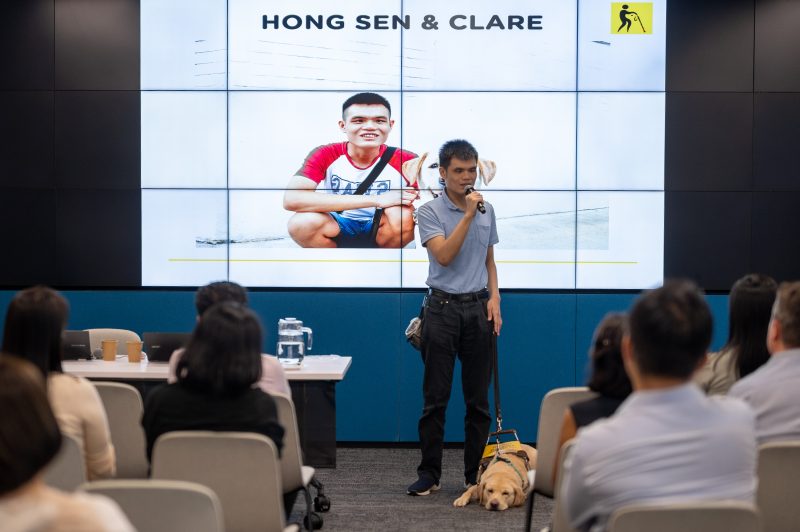 A GDS guide dog user stands at the front of the room and gives a sharing session, while his yellow Labrador guide dog peacefully sleeps beside him.