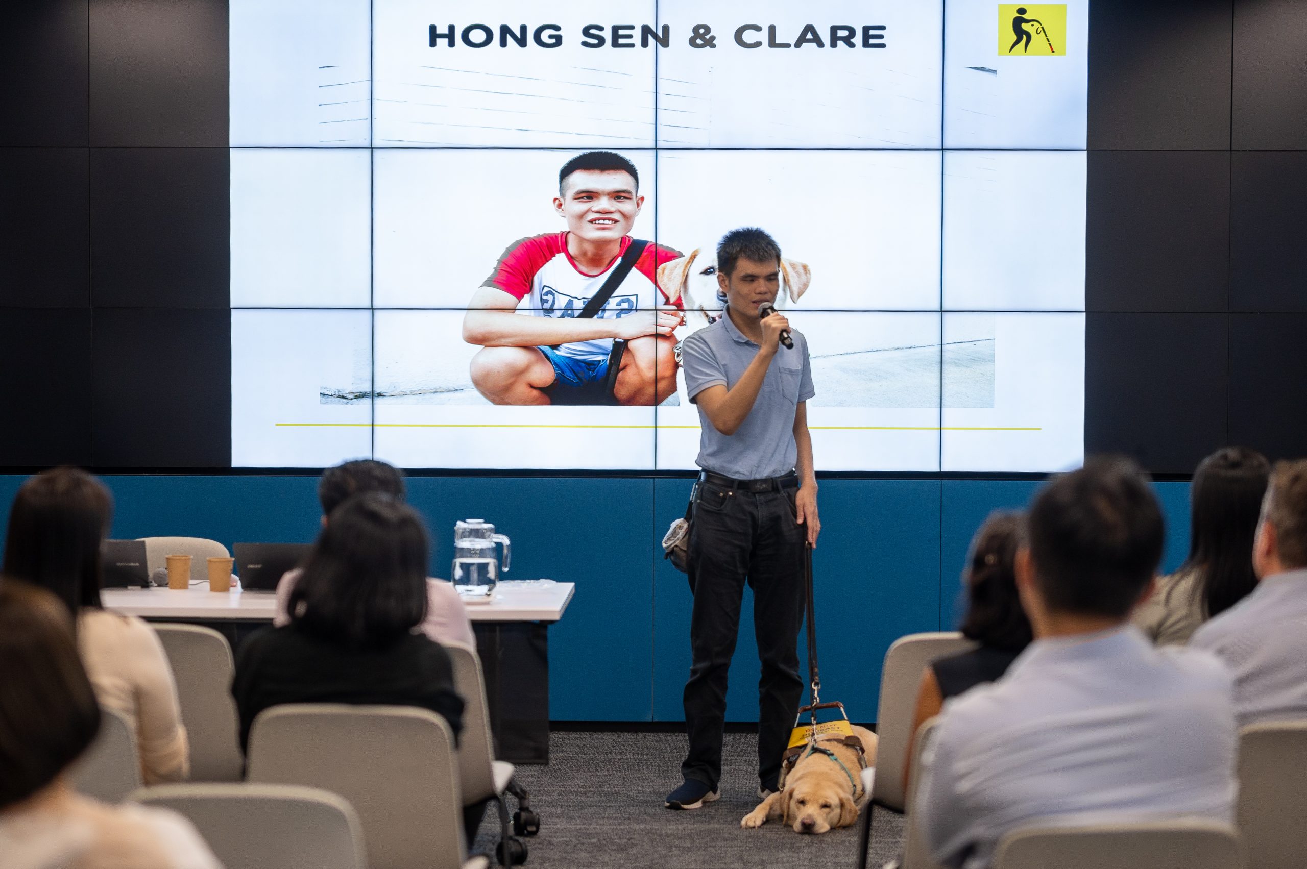A GDS guide dog user stands at the front of the room and gives a sharing session, while his yellow Labrador guide dog peacefully sleeps beside him.