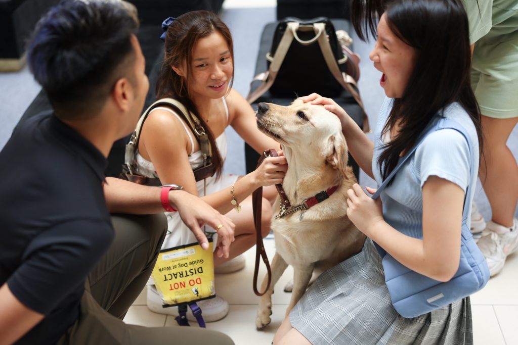 An event attendee expresses enthusiasm while petting Orinda, the guide dog, with Sophie, a guide dog user, squatting beside them. An event attendee expresses enthusiasm while petting Orinda, the guide dog, with Sophie, a guide dog user, squatting beside them.