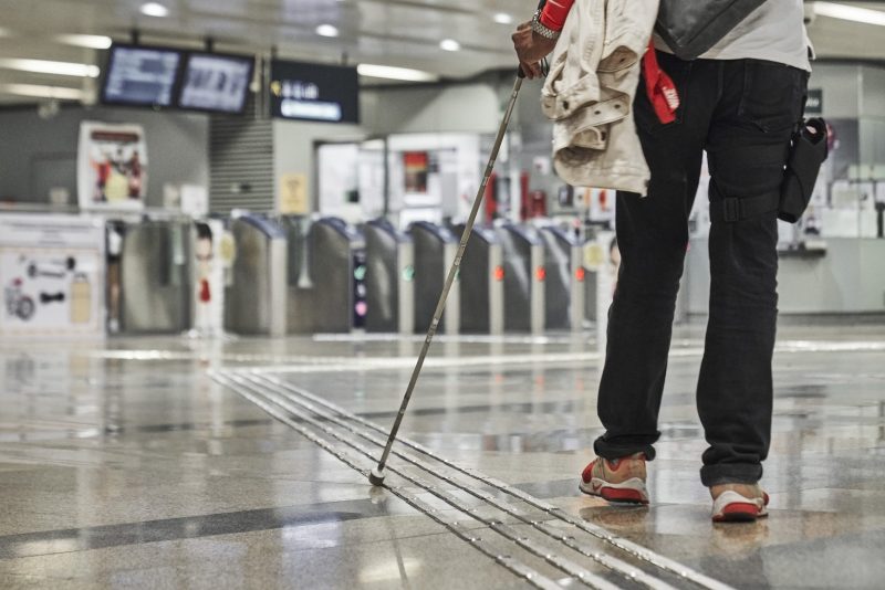 Close-up photo of a GDS client using a white cane to navigate tactile markers on an MRT platform.