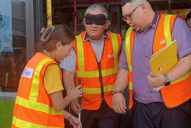 A bus captain is blindfolded and holding a cane while receiving sighted help from another bus captain to alight from the bus. A GDS staff member is assisting the pair in alighting. A collage of two photos. The left photo shows a bus captain who is blindfolded and holding a cane, receiving assistance from another bus captain as they alight from the bus. A GDS staff member is also helping them. The right photo features GDS staff smiling and posing for the camera with two guide dogs.