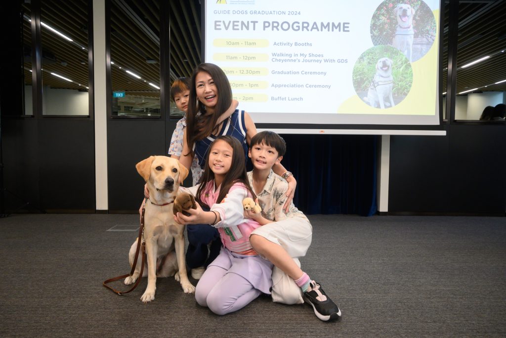 An event attendee poses for a group photo with a yellow Labrador guide dog and her three children. The children are holding small dog plushies in their hands. 