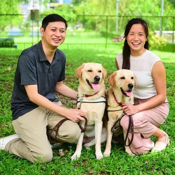 Sebastian and Sophie are kneeling on a patch of grass with their guide dogs, smiling and posing for a photo.