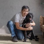 Alvin kneels with his left leg down and right leg up, lovingly hugging his black guide dog, Laurie, with both hands.