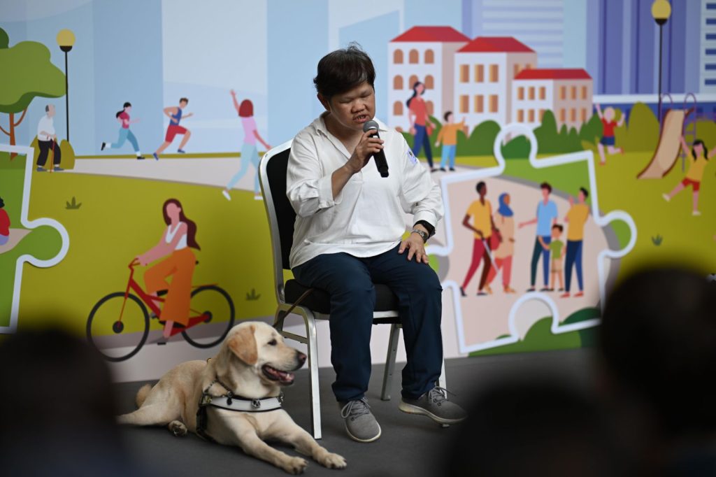 Guide dog user giving a presentation on stage, with her Yellow Labrador guide dog lying beside her.