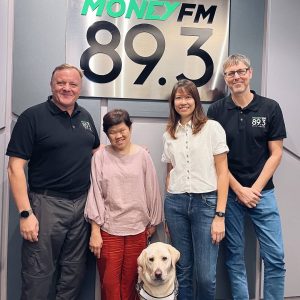 Lee Lee with her guide dog, Christina, and Money FM89.3 hosts Neil and Glenn posing together with a Money FM89.3 sign in the background.