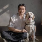 Thomas, a vision-impaired man, sits cross-legged with his white guide dog, Eve, by his left side, wrapping his arm around her affectionately.