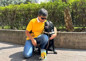 A guide dog user crouches next to his black Labrador guide dog in front of a Flag Day 2024 donation tin.