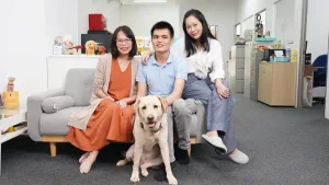 Two GDS staff members and a GDS guide dog user sitting on a sofa, posing for the camera with a yellow Labrador guide dog.