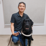 Guide dog user, Manabu, crouching next to his black guide dog, Momo.