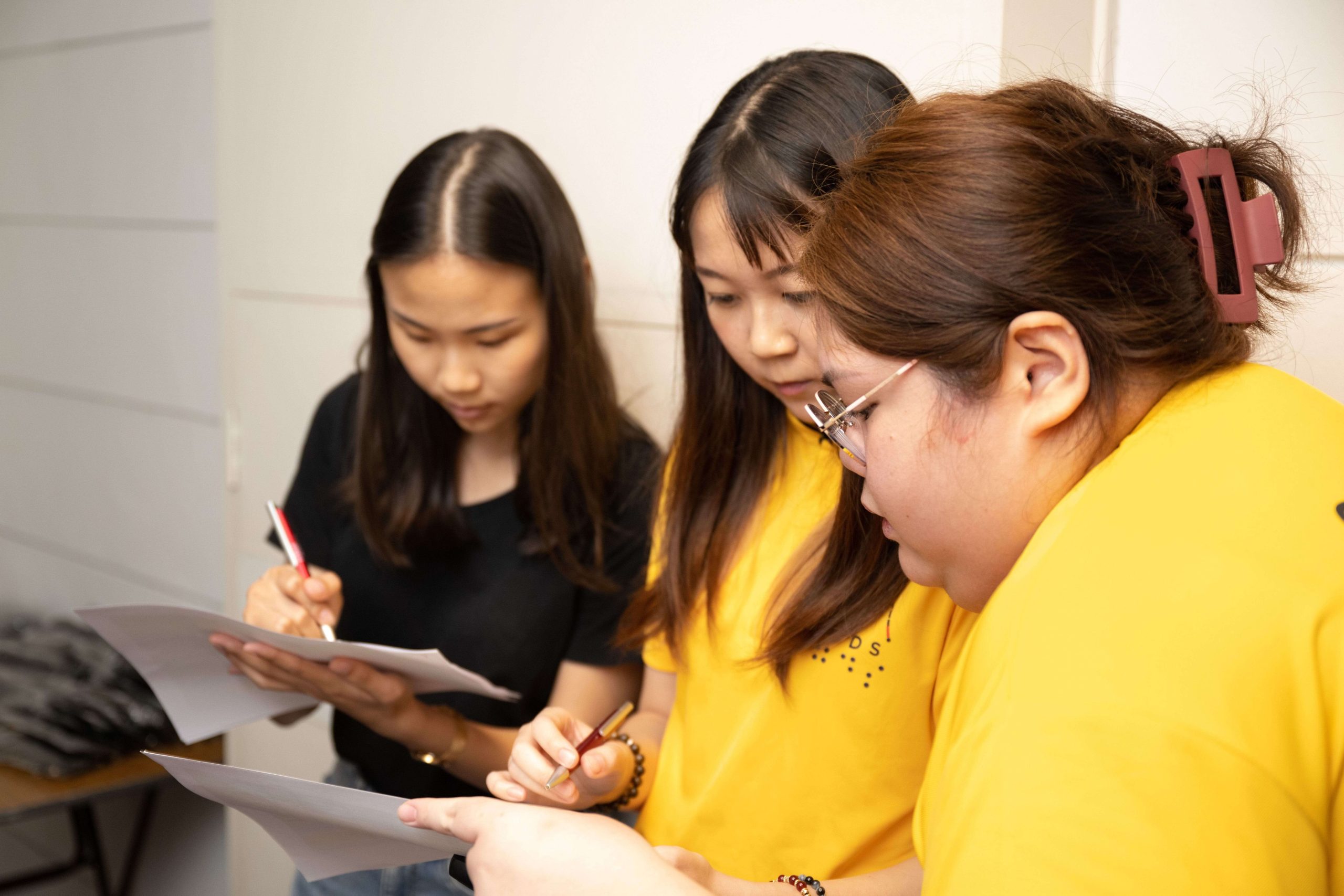 Three young women standing indoors, holding papers and pens while focusing on writing or discussing documents together.