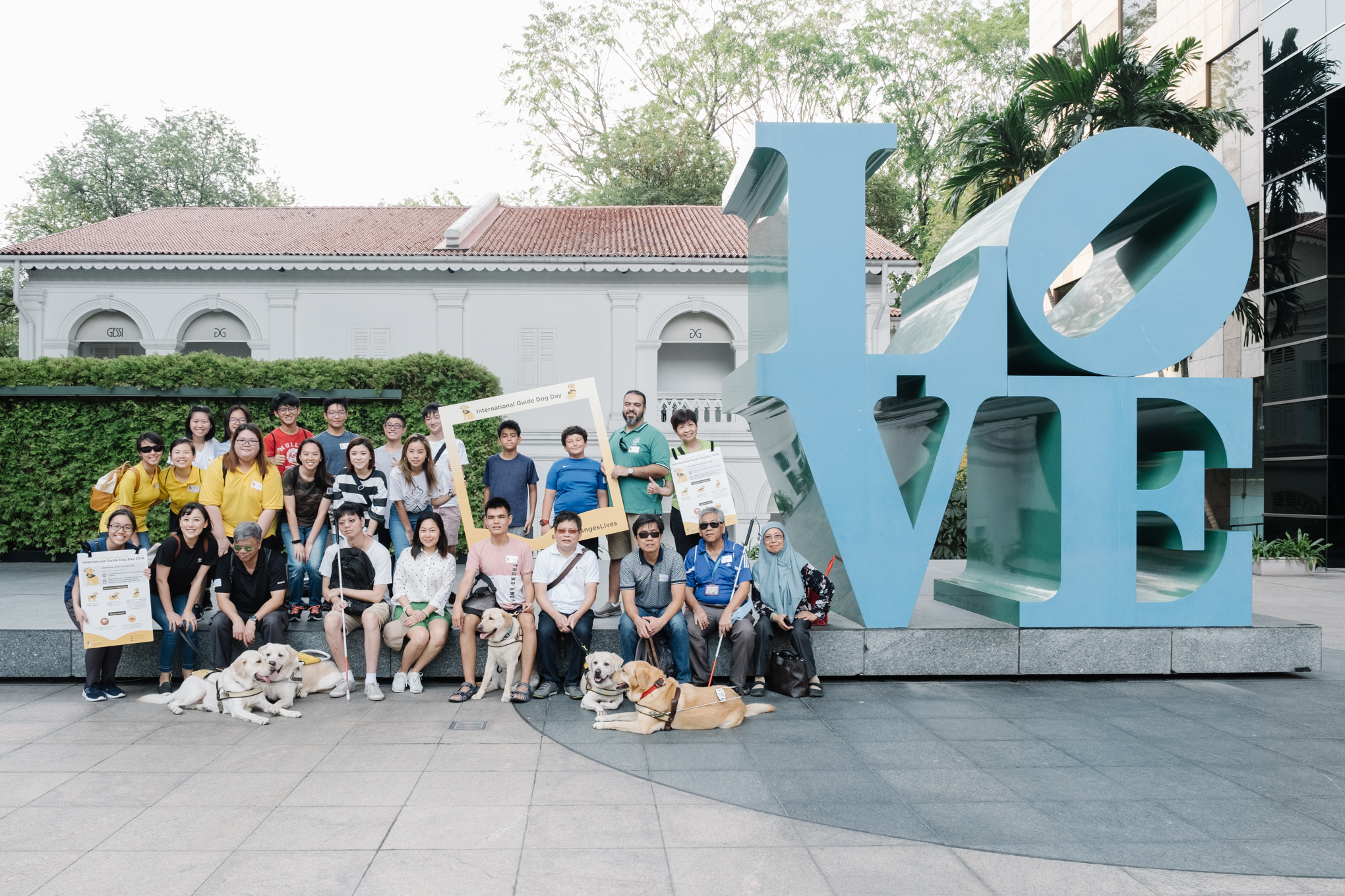 A large group of people, including guide dog users and volunteers, pose together in front of a giant blue "LOVE" sculpture outdoors. Several guide dogs lie or sit at the front, and participants hold yellow posters and frames celebrating International Guide Dog Day.